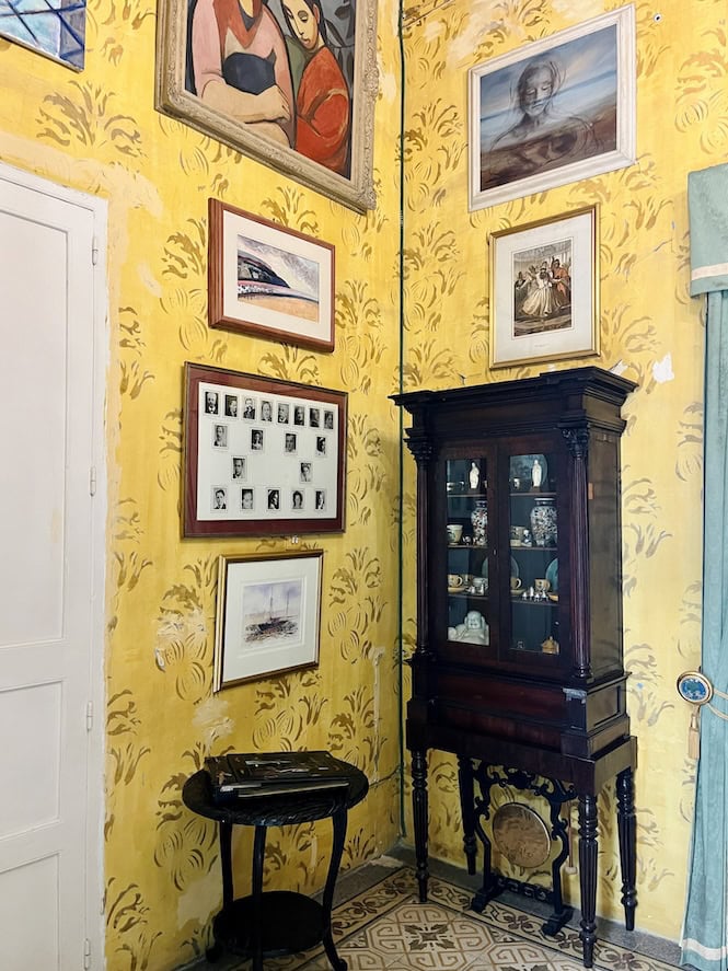 Corner of an ornate yellow room with antique furniture, multiple framed artworks, and a display cabinet, showcasing the Chinese Room at Casa Rocca Piccola in Valletta, Malta.