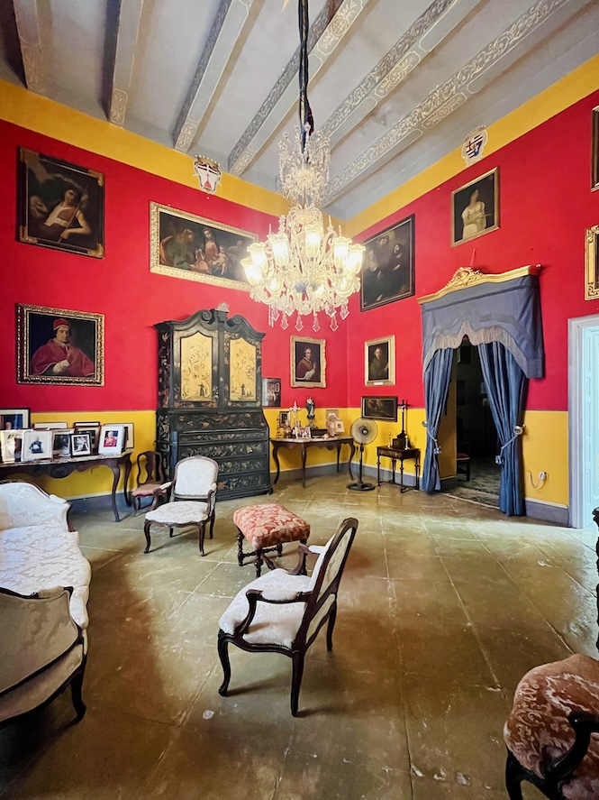 Opulent Sala Grande room with red and yellow walls, ornate chandelier, antique furniture, and numerous paintings, showcasing Maltese and European historical decor at Casa Rocca Piccola.