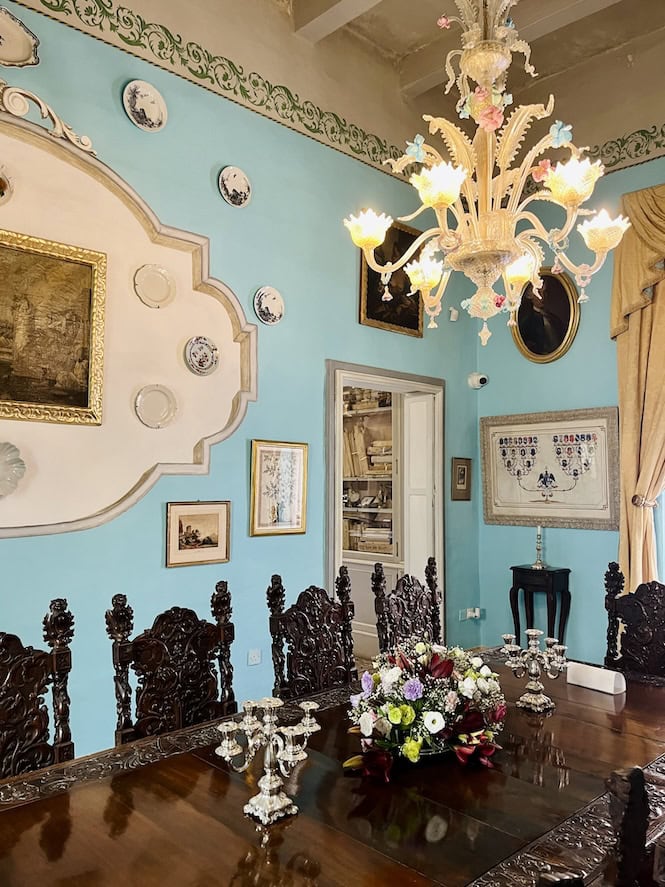 Elegant dining room with light blue walls, ornate chandelier, dark wooden carved chairs, and various framed artworks.