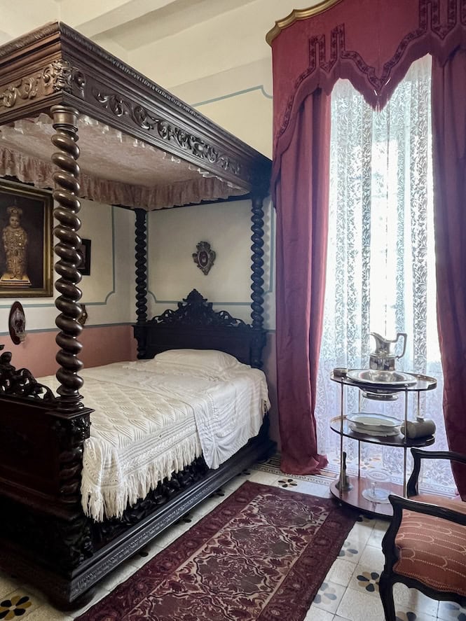 Ornate four-poster bed with dark wooden frame and white bedding. Rich burgundy curtains and decorative rugs adorn the room.