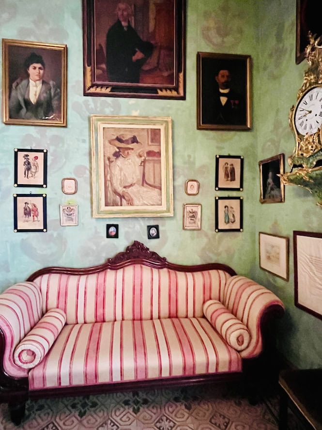 Room with pale green walls covered in framed portraits and artwork. A pink and white striped sofa sits beneath the gallery wall.