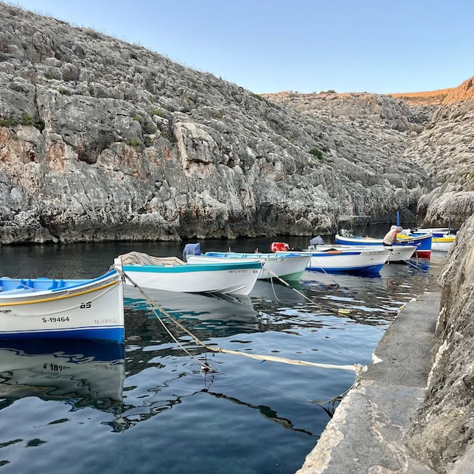 A stunning Blue Grotto cove in Malta with clear blue water and parked boats, demonstrating the warm weather and beauty of Malta in October.
