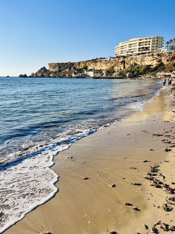 Golden Bay, Malta, is renowned for its beautiful sandy beach and crystal-clear waters, making it one of the best areas to stay in Malta for beaches. Visible in the image is the Radisson Blu Resort & Spa, Golden Sands, perched on a cliff overlooking the bay. The hotel offers luxurious accommodations with stunning sea views and easy access to the beach.