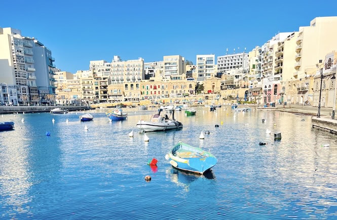 Spinola Bay in St. Julian's, Malta, is a vibrant and scenic spot known for its excellent restaurants and lively nightlife. The bay is lined with modern buildings, restaurants, and cafes, offering a variety of dining options and nightlife venues. Small boats are moored in the calm waters, adding to the picturesque charm of this popular area.