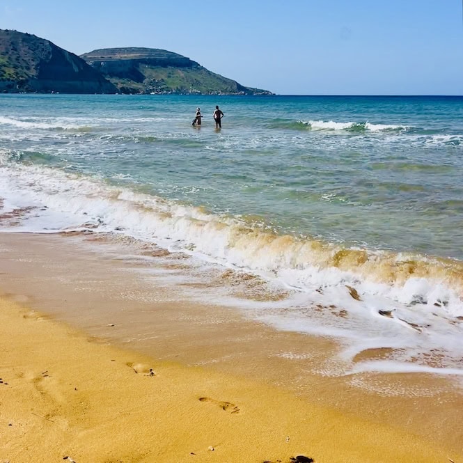 Stunning Ramla Beach, a popular tourist attraction in Gozo, Malta.
Golden sand meets crystal-clear turquoise waters of the Mediterranean Sea.
Two people wading in gentle waves with scenic hills in the background.