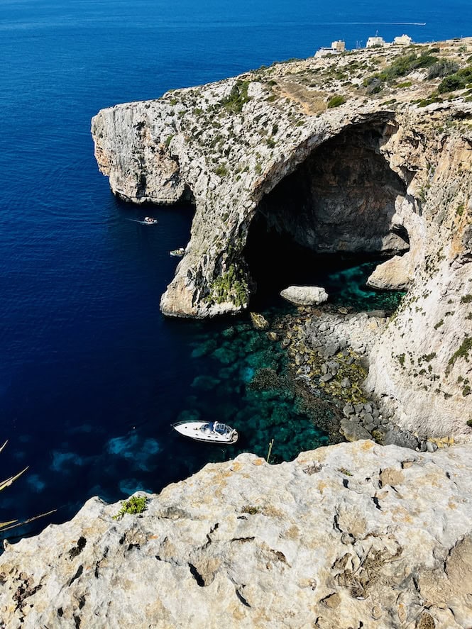 A scenic view of the Blue Grotto in Malta, showcasing a large rock arch over the deep blue sea with boats navigating the clear waters below. This popular tourist attraction is renowned for its vibrant underwater landscapes and dramatic coastal formations.