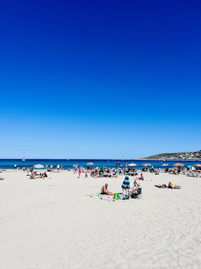 Ghadira Bay, Malta, showcases a lively beach scene with sunbathers and colorful umbrellas dotting the sandy shore. This popular tourist attraction is known for its clear blue waters and family-friendly atmosphere.