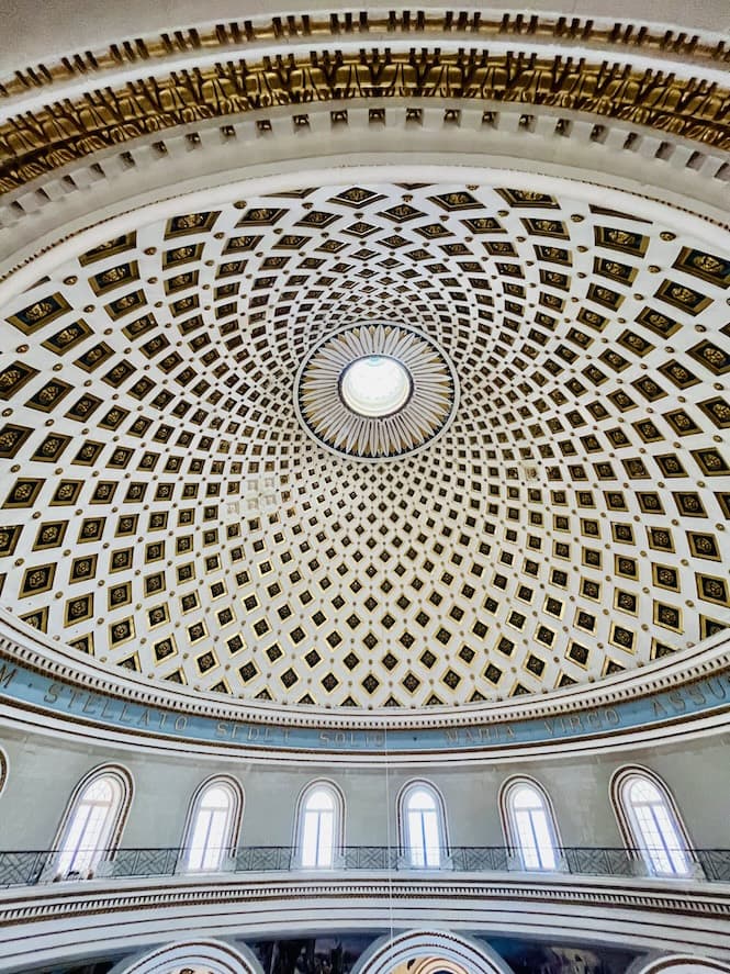 The image shows an impressive domed ceiling of the Mosta Rotunda with intricate geometric patterns and numbered panels radiating out from a central point. This appears to be the interior of a grand building, likely a church or cathedral. Mosta Rotunda is a religious site that appeals to tourists interested in architecture and cultural heritage.