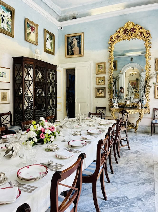 The image shows the summer dining room of Casa Rocca Piccola, a 16th-century palace in Valletta, Malta. It features a long table set for a formal meal, walls adorned with paintings, and a large ornate mirror. The elegant room, with its light blue ceiling and antique furnishings, offers visitors a glimpse into the lifestyle of Maltese nobility, serving as a living museum of the island's cultural heritage.