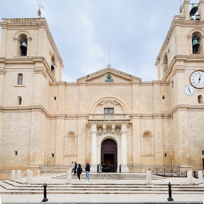 The photo displays the front of St. John's Co-Cathedral in Valletta, Malta. It may seem simple at first glance, but this popular tourist spot is home to breathtaking Caravaggio paintings. The Co-Cathedral, constructed from limestone, features a clock on its right side.