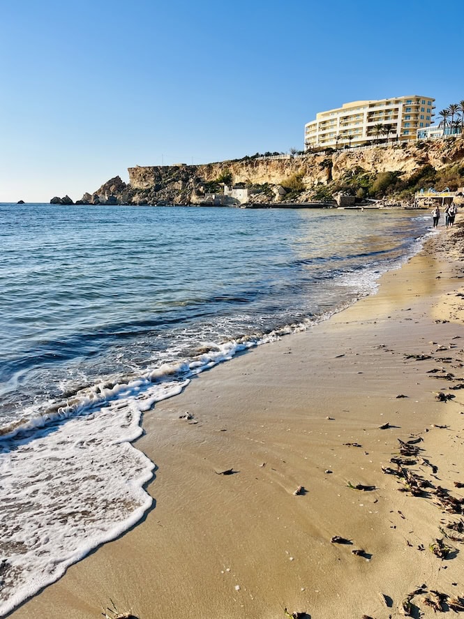 Golden Bay, Malta, is depicted here with its pristine sandy beach meeting the gentle waves of the Mediterranean Sea. A luxury hotel sits atop the rugged cliffs, offering stunning views of this popular tourist attraction.