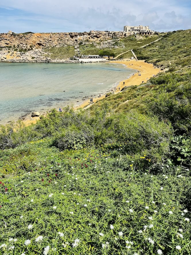 Ghajn Tuffieha, Malta, showcases its picturesque beach and tranquil bay, surrounded by lush greenery and rocky cliffs. Visitors can be seen enjoying the scenic landscape of this popular tourist attraction.