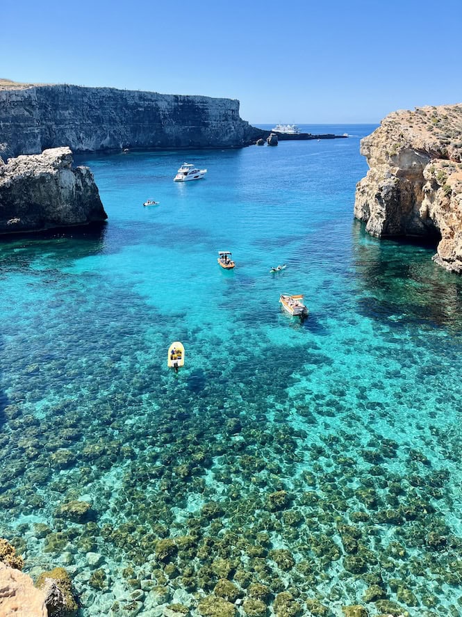 Crystal Lagoon, a stunning tourist attraction in Malta. Turquoise waters surrounded by towering limestone cliffs. Small boats and yachts scattered in the clear, shallow bay. Visible coral reefs beneath the surface create a picturesque Mediterranean seascape.