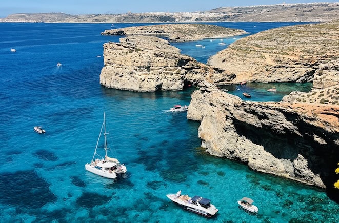 Aerial view of a stunning crystal lagoon surrounded by rugged cliffs in Malta, a popular tourist attraction. The clear turquoise waters are dotted with boats and yachts, showcasing the area's appeal for maritime activities and sightseeing.