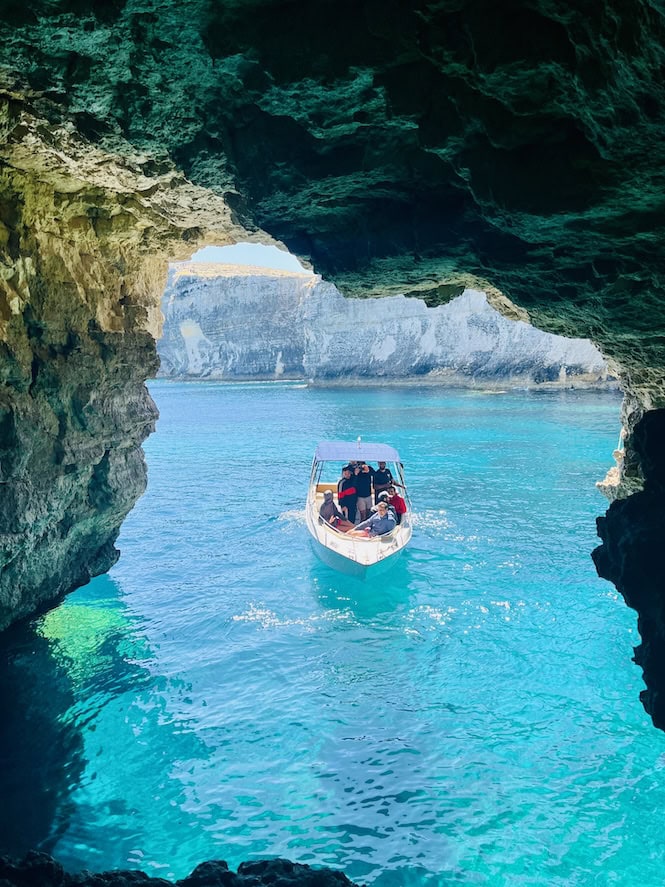 Santa Maria Caves, Comino: Breathtaking view of turquoise waters through a natural rock arch. Small boat with tourists exploring the cave entrance, surrounded by dramatic limestone formations and crystal-clear Mediterranean Sea.