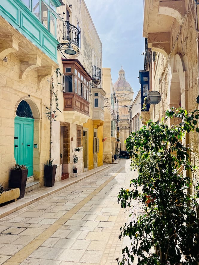 The streets in Three Cities, featuring brightly colored windows and doors, adding a lively contrast to the ancient stone facades. A magnificent dome is seen in a distant, symbolizing Malta's rich cultural heritage. These historic streets, with their unique architecture and vibrant details, are a must-see Malta's tourist attraction.