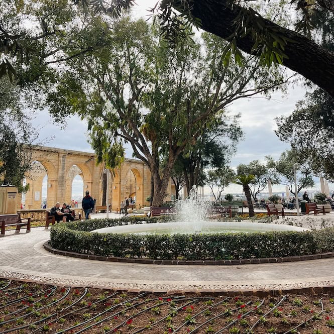 Scenic view of Upper Barrakka Gardens in Malta, featuring a central fountain, arched colonnade, and lush trees overlooking the sea, a popular tourist attraction.