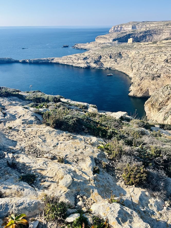 Panoramic coastal view of Dwejra in Gozo, Malta. Rugged limestone cliffs overlooking the deep blue Mediterranean Sea. Ancient watchtower visible in the distance.