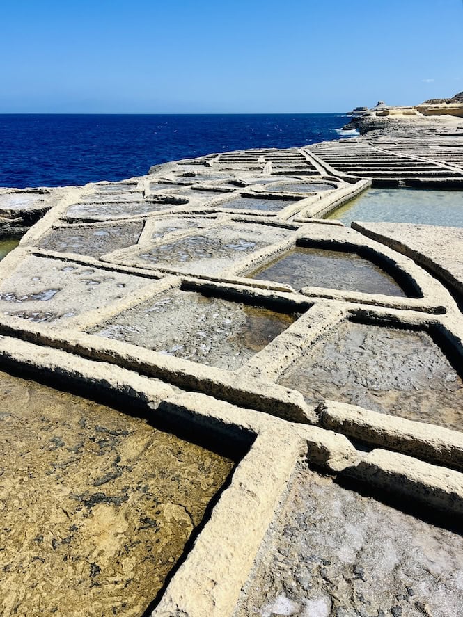 Ancient salt pans carved into coastal rocks in Malta. Geometric pattern of shallow pools filled with seawater against deep blue Mediterranean Sea. 