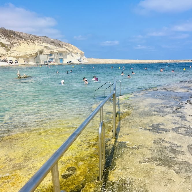 Swimmers enjoying the shallow waters of Xwejni Bay in Gozo with safety railings visible