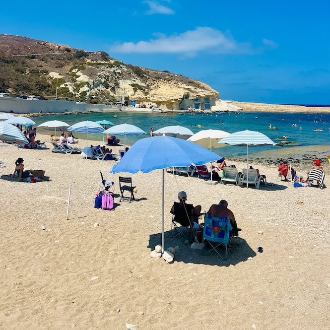 Beachgoers relaxing under umbrellas on the sandy shore of Xwejni Bay in Gozo