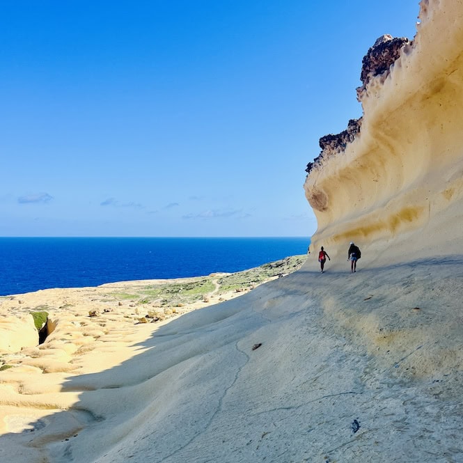 Two hikers walking along a rocky coastal path with stunning views of the Mediterranean Sea, showcasing a must-do hiking experience in Malta.