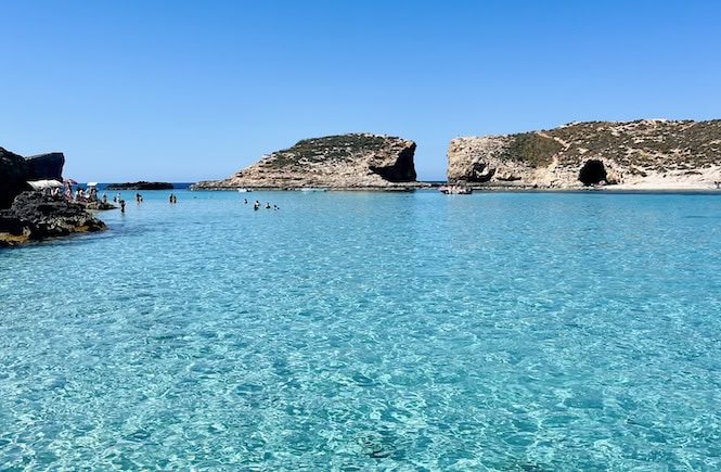 Crystal clear waters of the Blue Lagoon in Comino, Malta, with people swimming and rocky islands in the background, showcasing a must-do experience in Malta.