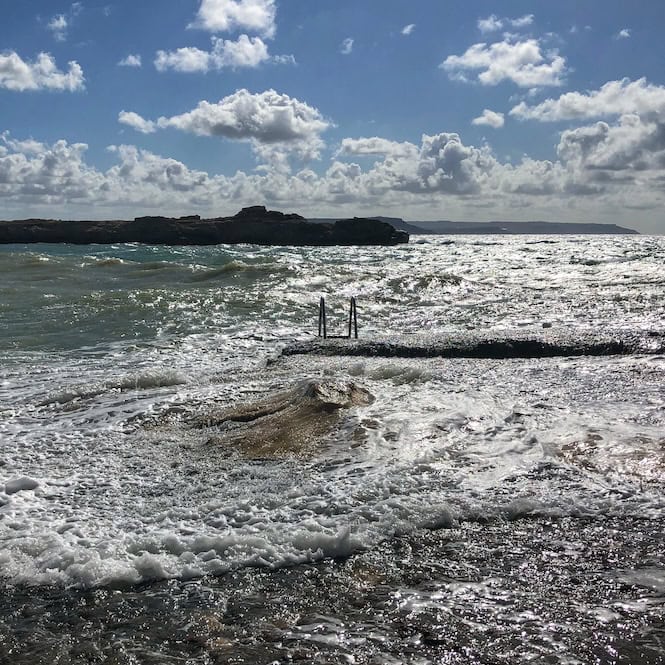 Rough sea waters crashing against a submerged ladder and platform on a rocky shoreline in Malta under a partly cloudy sky – a reminder why you shouldn’t go close to rough water.