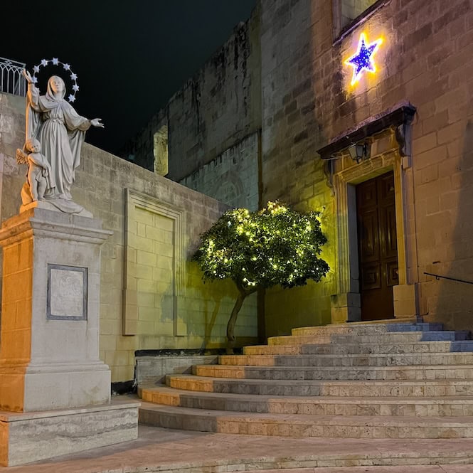 A serene night view of a Maltese church decorated for Christmas, featuring a statue of the Virgin Mary, a lit tree, and a glowing star above the entrance, showcasing Malta's festive spirit and religious traditions.