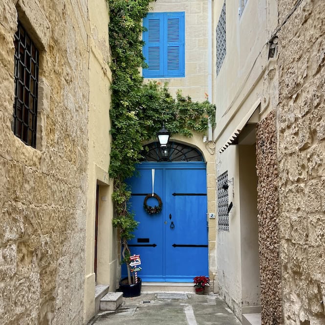 A picturesque blue door in a narrow Maltese alley, adorned with a simple Christmas wreath and festive plants, highlighting the cozy holiday charm of Malta’s traditional streets.