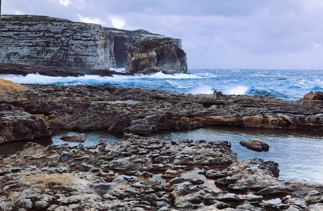 Rocky coastline in Malta with dramatic waves crashing against rugged cliffs under a cloudy sky – a striking view of the island's natural beauty and powerful sea.