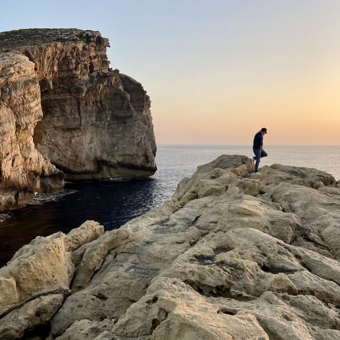 Person standing on a rocky cliff overlooking the calm sea at sunset in Malta, near towering limestone formations – a peaceful yet potentially risky spot to explore.