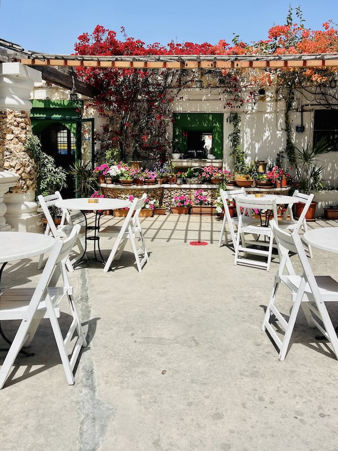 A Mediterranean courtyard cafe with white folding chairs and round tables arranged on a concrete patio. The space features vibrant red bougainvillea climbing over a wooden pergola, green-framed windows on whitewashed walls, and stone accents. Various potted plants and pink flowers decorate the perimeter, creating a charming garden atmosphere under bright blue skies.