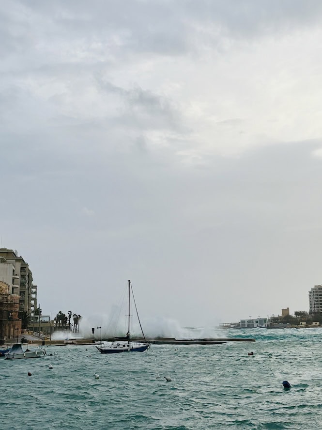 Dramatic January storm waves crashing against St Julian's promenade in Malta, with moored boats battling rough Mediterranean seas under cloudy skies