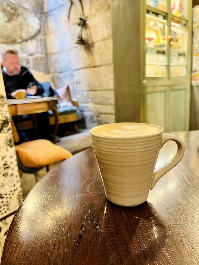 A beige ceramic coffee cup with latte art sits on a dark wooden table inside a cozy café with limestone walls and vintage green cabinets in the background. A blurred patron sits at a distant table.