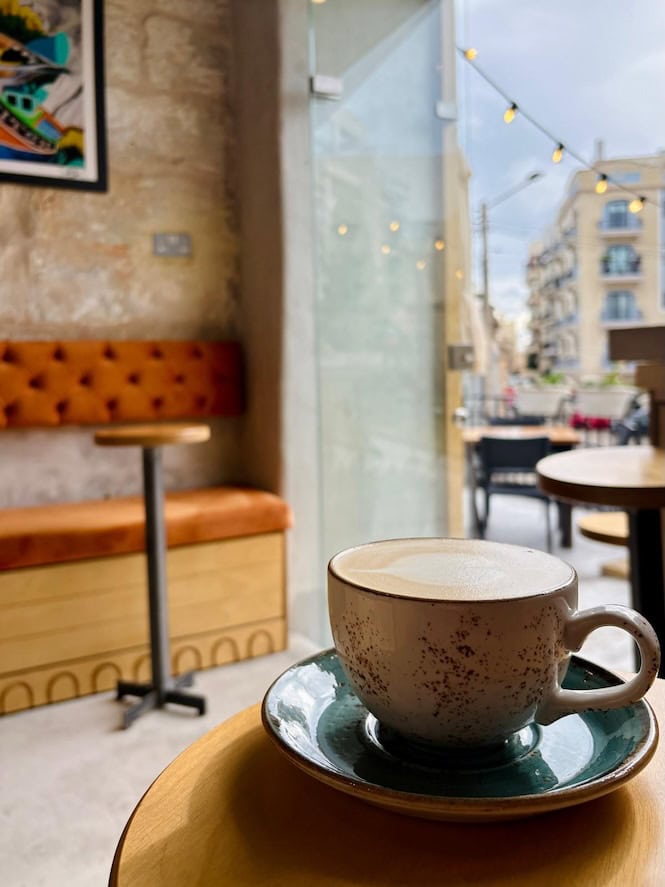 Cappuccino served in a ceramic cup at Brillace café in Malta, with cozy seating and city street view in the background.