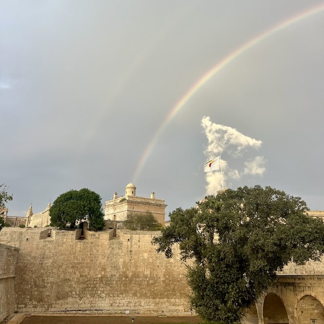 A colorful rainbow arcs above the historic city of valletta in Malta, with its iconic limestone walls and Maltese flag fluttering in the breeze after rainfall.