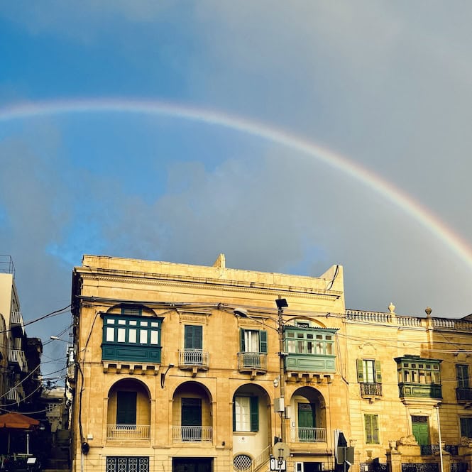 A vibrant rainbow arches across a partly cloudy sky above traditional Maltese limestone buildings with colorful enclosed wooden balconies, following a rain shower in Malta.