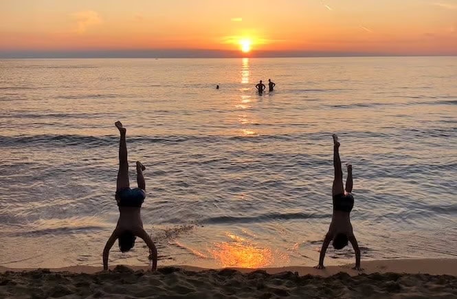 Two children doing handstands on the beach at sunset, with people swimming in the calm sea at Golden Bay, Malta.