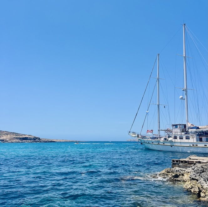 Sailing yacht anchored in crystal-clear waters on a bright summer day in Malta, near rocky coastline and open sea.