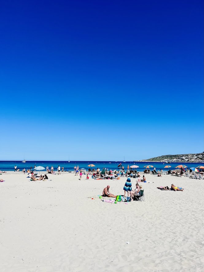 People relaxing and sunbathing on the wide sandy shore of Mellieħa Beach under a clear blue sky, highlighting one of the best free things to do in Malta.